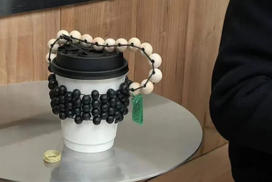 A beaded cup carrier with a handle on a takeaway coffee cup.