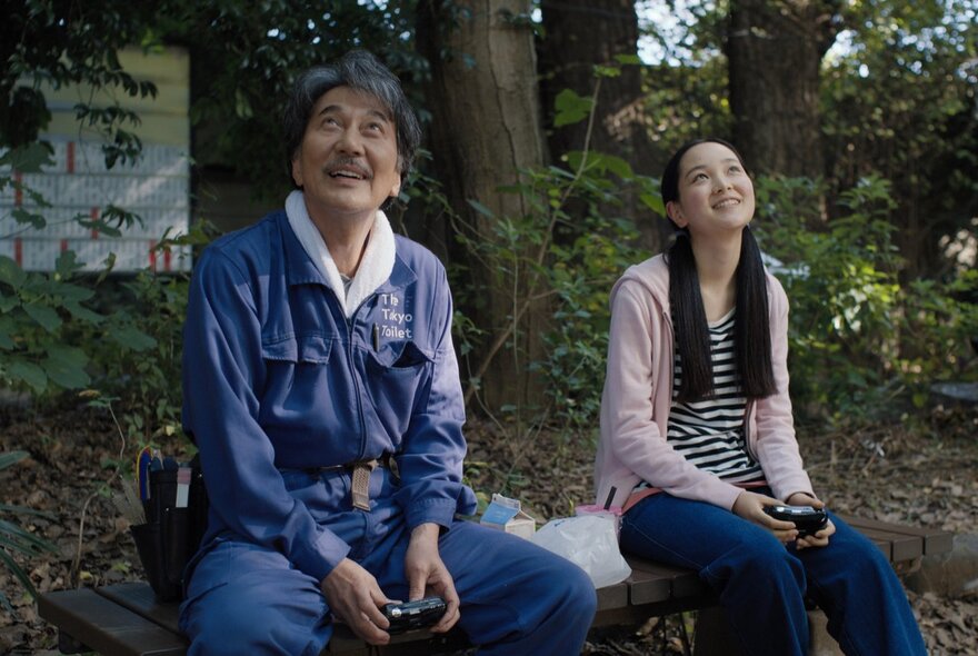 A man in a blue utility uniform and a young woman in a striped shirt and pink cardigan sit on a wooden bench outdoors, both looking up at the trees and smiling.The image is a still from the film 'Perfect Days'.