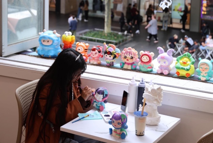 A woman painting a plaster figurine at a table in front of an open window overlooking the street, with completed figurines lined up on the window sill.