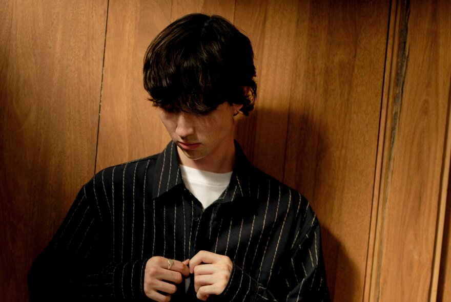 A male fashion model buttoning up a dark long sleeved shirt, leaning against a wood panelled wall.