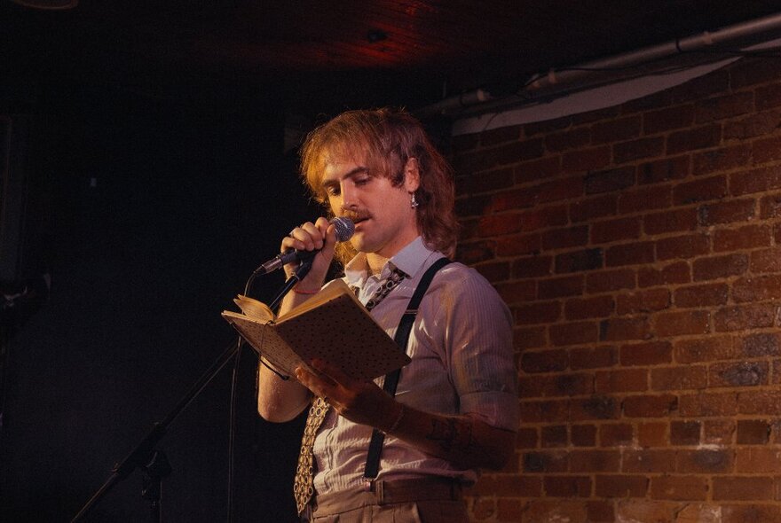 Man standing at a microphone reading from a book, wearing braces, in a dimly lit brick-walled venue.