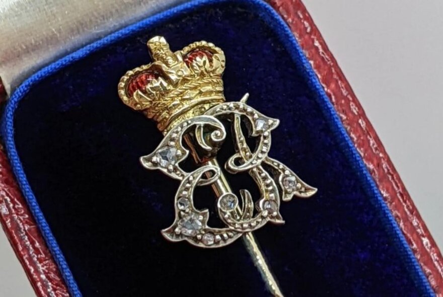 A piece of royal presentation jewellery featuring a crown and the ER insignia of Queen Elizabeth II, resting on a blue velvet cushion inside a jewellery box.