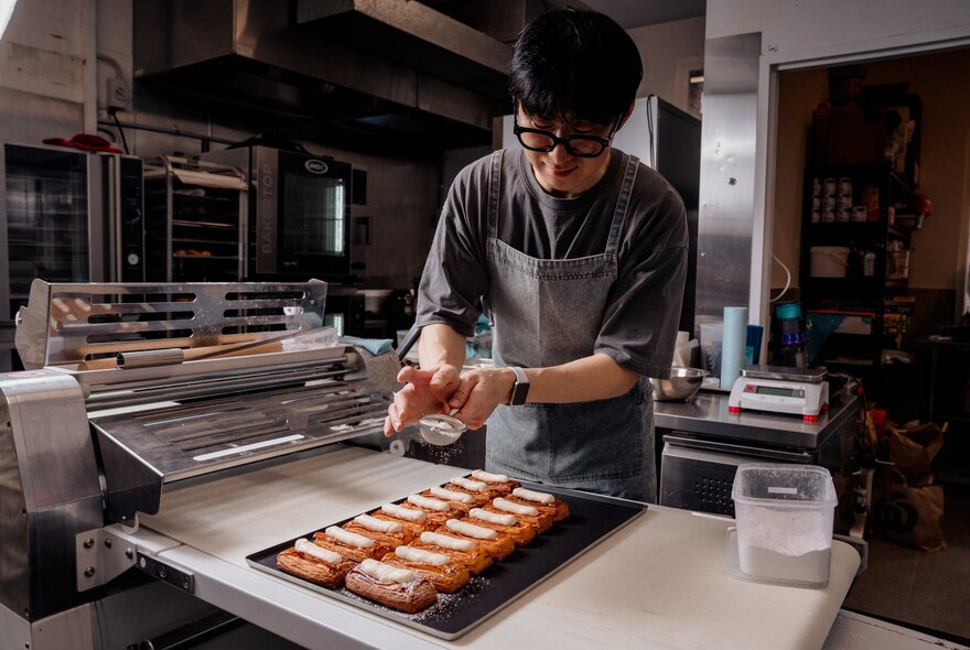 Baker adding icing sugar to rows of baked pastries.