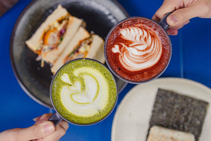 Two hands bringing two cups of hot beverages together in a clinking motion above a plate of food that rests on the table below.