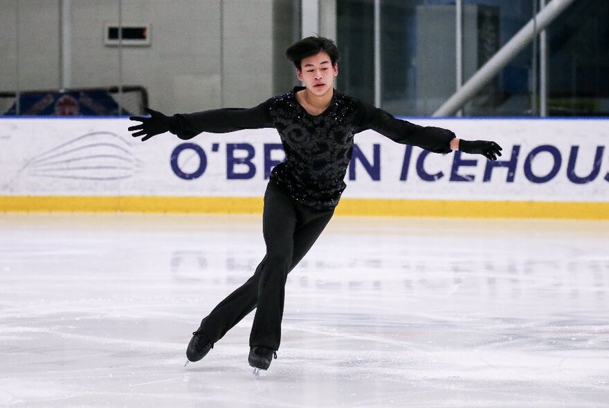 A male figure skater performing on ice in a black suit and gloves. 