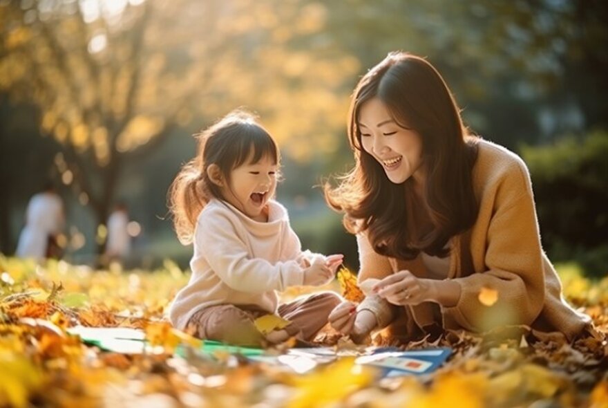 Mother and child seated on autumn leaves in dappled sunshine playing with books.