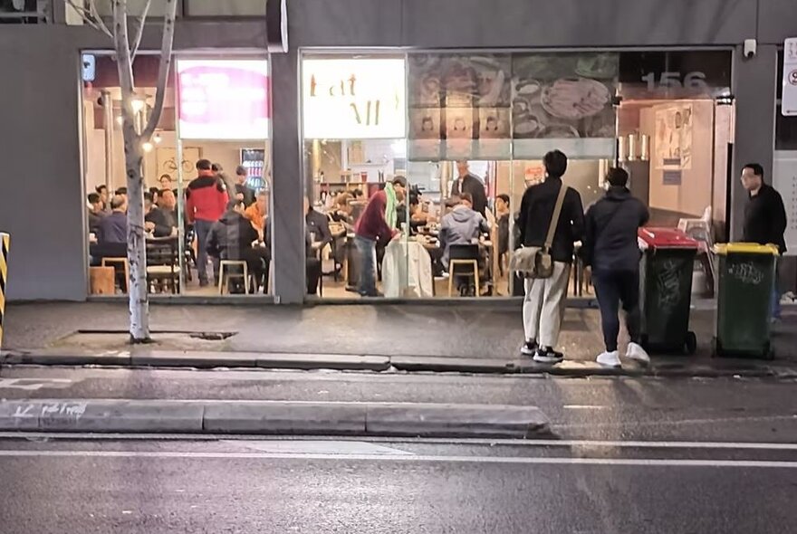 The exterior of restaurant Eat All  with people waiting outside near wheelie bins and lots of people inside at tables and walking around. 