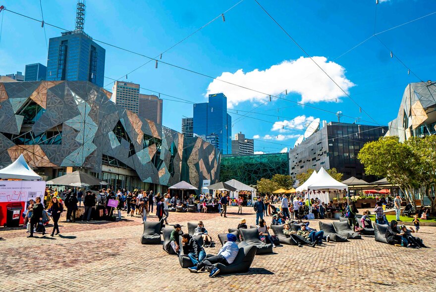 A crowd of people are gathered at a festival in Fed Square. Some of the people are sitting on beanbags.