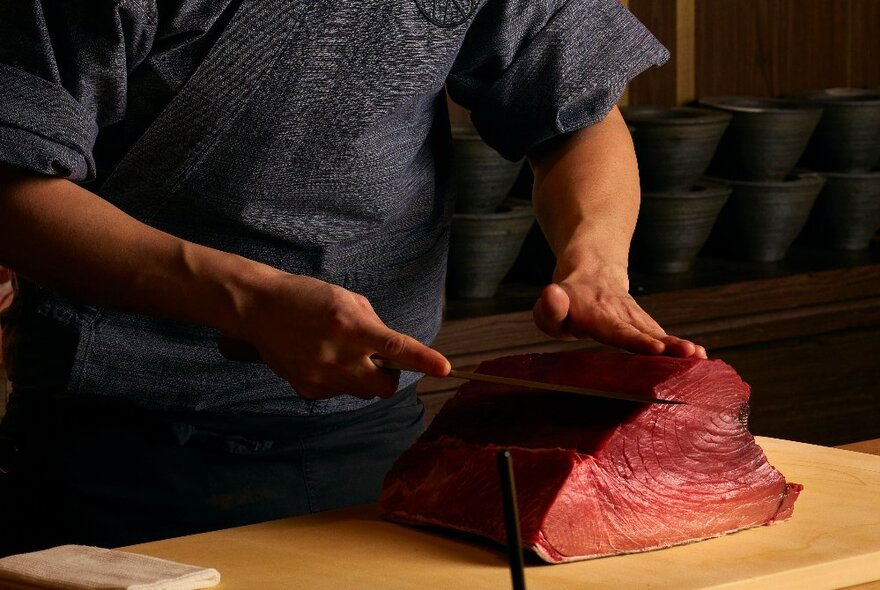 A chef expertly slicing into a large portion of red bellied tuna on a wooden chopping board.