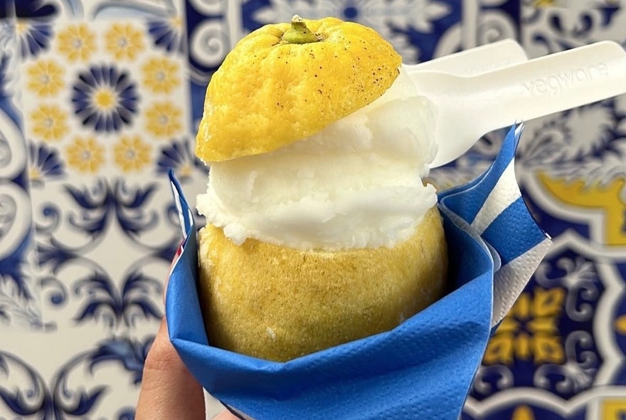 A lemon sorbet with a small spoon sticking out of it, served inside a hollowed out real lemon, wrapped in a blue and white striped napkin, against a blue, yellow and white patterned tile background.