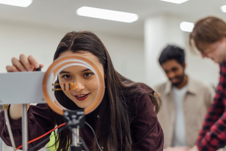 A young woman peering into an orange circle in a science lab with two young men behind her. 