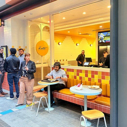 The yellow storefront and outdoor seating area of Kantin Indonesian restaurant, with people inside ordering food and queued near the door.