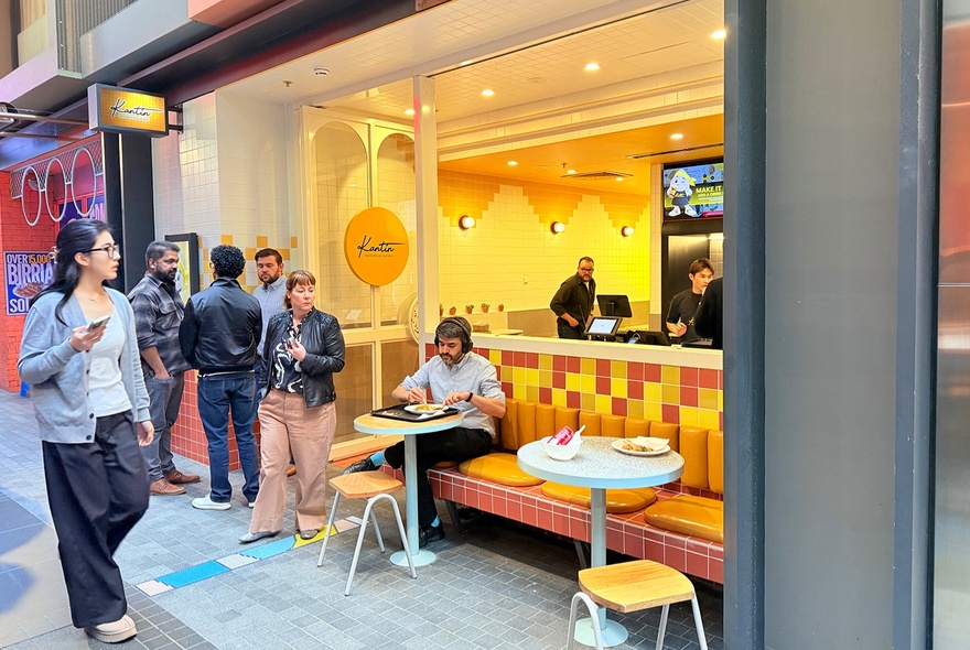 The yellow storefront and outdoor seating area of Kantin Indonesian restaurant, with people inside ordering food and queued near the door.