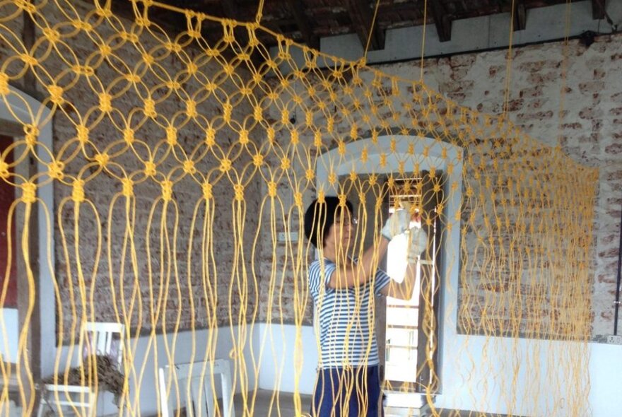 Person working on a large macrame artwork project in a brick-lined room.