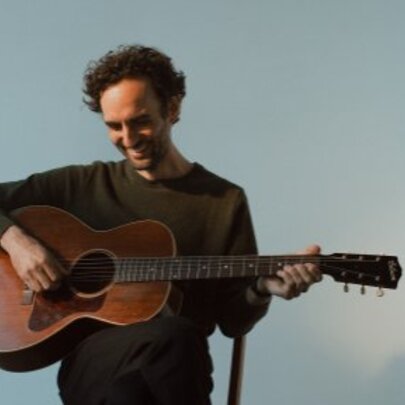 Guitar virtuoso and composer Julian Lage smiling and looking downwards while strumming his guitar. 