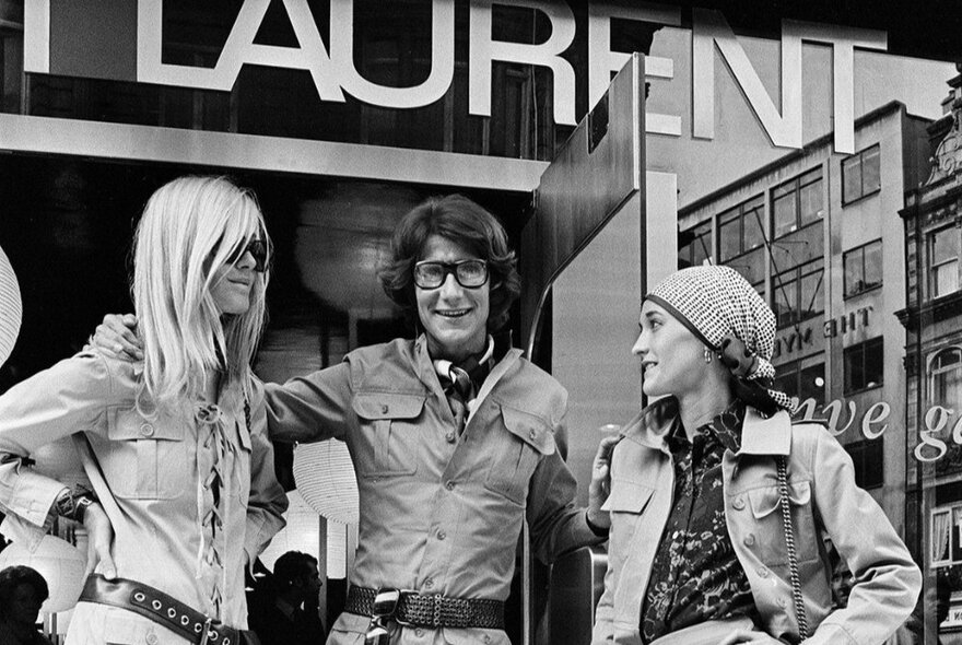 A black and white image of two models and a designer ourside of Yves St Laurent. 