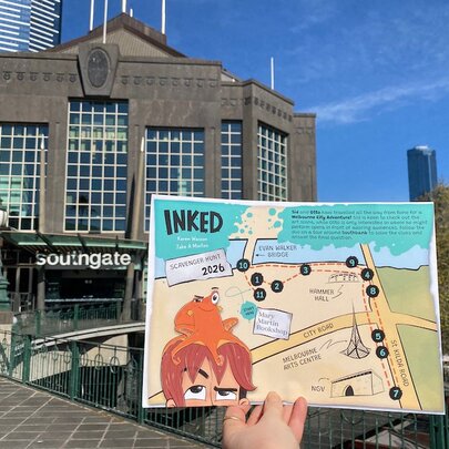 A person holding an 'Inked' scavenger map in front of the Southgate Melbourne complex in Southbank, with blue sky background.