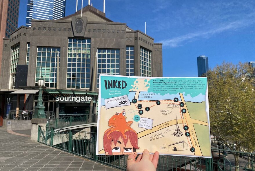 A person holding an 'Inked' scavenger map in front of the Southgate Melbourne complex in Southbank, with blue sky background.