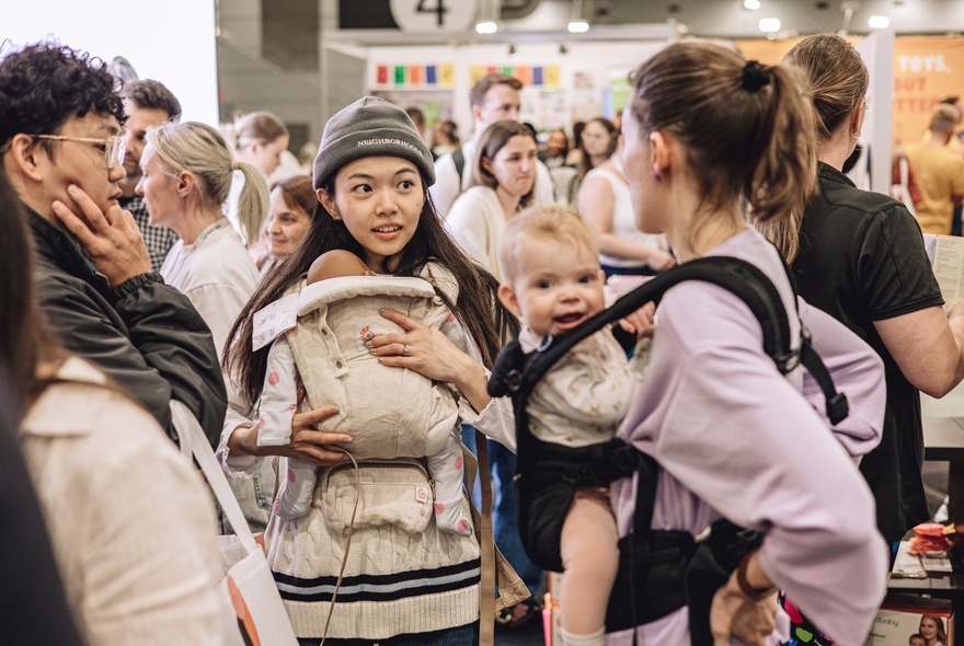 Two mothers carrying their young babies in carriers on their chest, talking to each other at The Pregnancy Babies & Children's Expo.