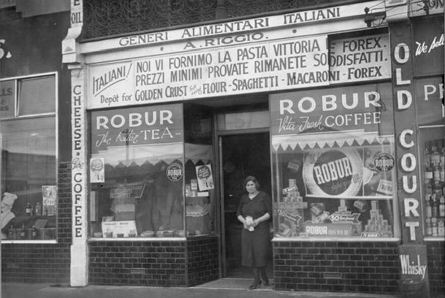 Vintage photo of a person standing in the doorway of an Italian delicatessen with multiple signs advertising produce and window signage for Robur tea.