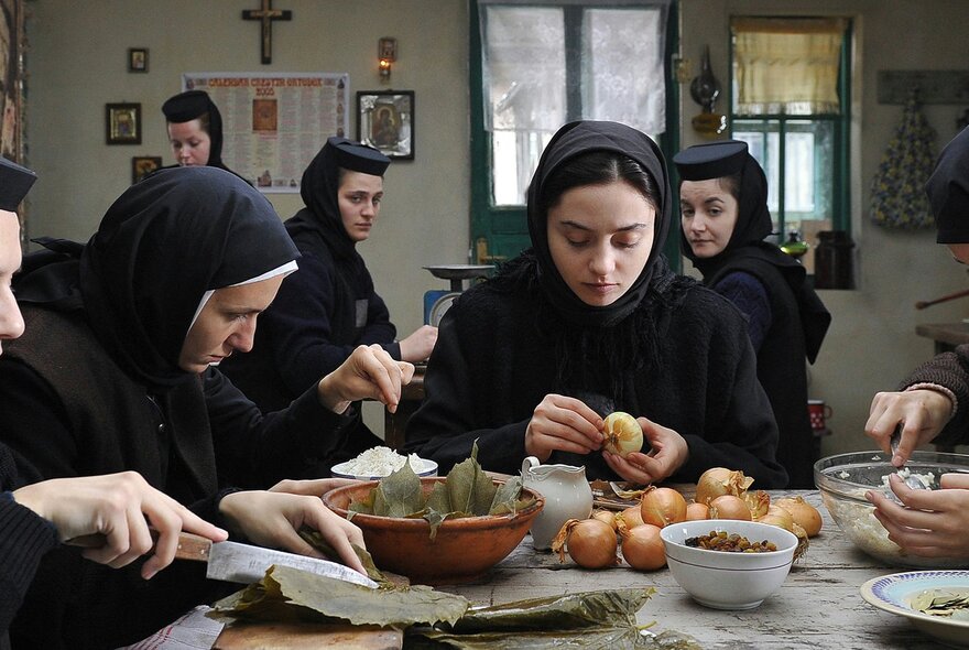 A still image from a film showing nuns in black habits all preparing food, one peeling onions. 