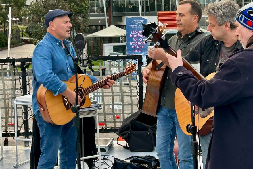 A group of musicians performing live with acoustic guitars on an outdoor stage.