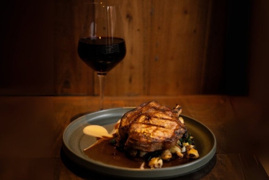 A moody image of a steak on a plate with sauce and a glass of red wine against a timber background. 