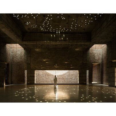 A lone person standing at the far end of a vast room dotted with lights, looking out a wide window towards an external brick facade.