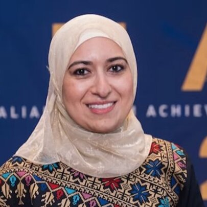 Dr Bushra Othman, wearing a pale head covering and a patterned top, she is standing in front of a branded wall.
