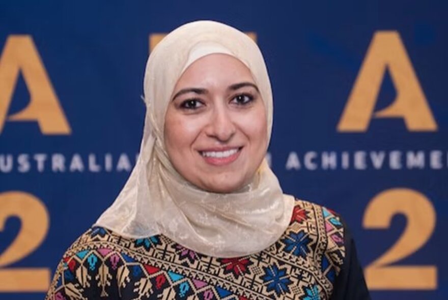 Dr Bushra Othman, wearing a pale head covering and a patterned top, she is standing in front of a branded wall.