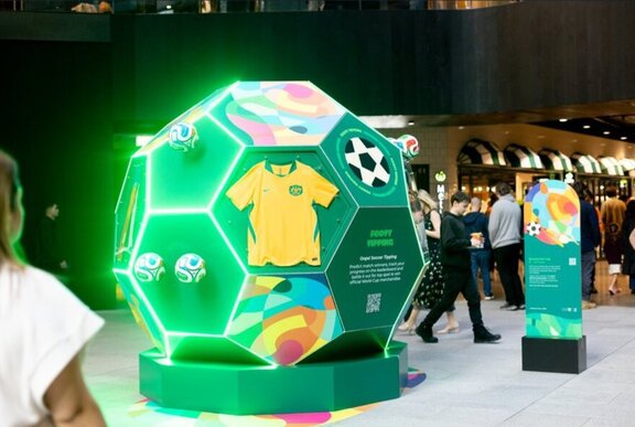 A large pop-up green soccer ball structure in the main retail plaza at Collins Square, covered with merchandise and signage, with pedestrians walking past.