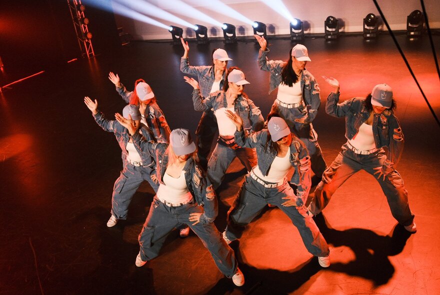 Seen from above, a group of eight dancers in matching denim jackets and pants, white tops, and caps perform hip hop on a stage with dramatic red and white lighting. 