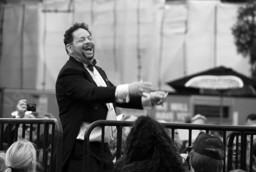 A male opera singer in a tuxedo, singing in an intimate setting with three people looking on.
