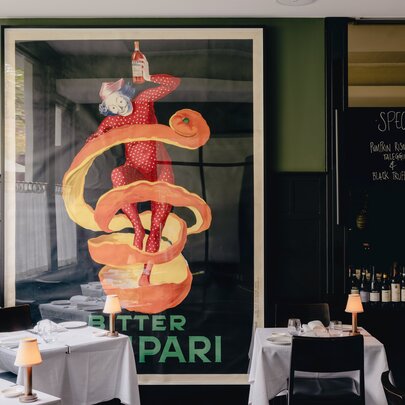 The dining room interior of Cafe Florentino restaurant on Bourke Street with a billboard-size Campari poster on the wall, tables covered in white linen and dark dining chairs.