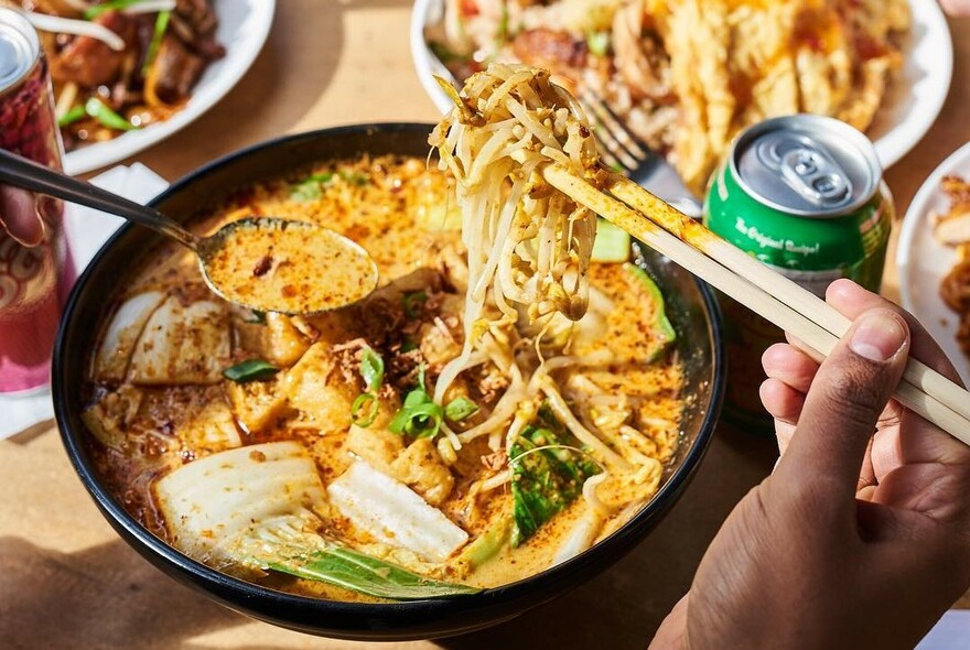 Hands with spoon and chopsticks selecting sauce and noodles from a black dish.