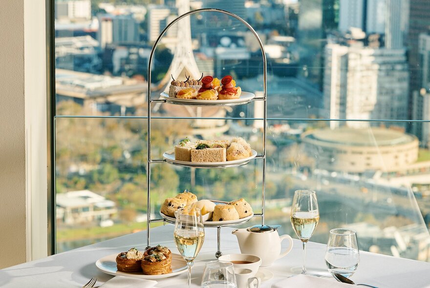 Looking at a table set for high tea with a 3-tier cake stand, in front of a window that looks out over the Melbourne skyline. 