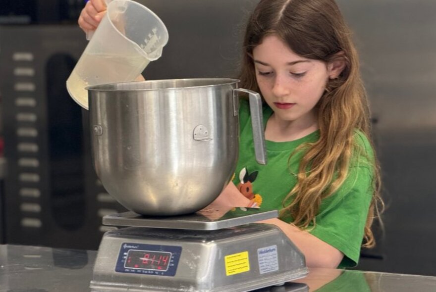 A child pouring liquid into a mixing bowl that is resting on kitchen scales on a stainless steel work bench.