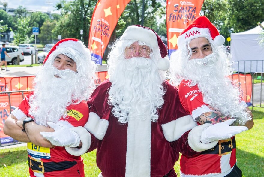 Three people dressed as Santa, two of them wearing running race bibs, standing outside in a a park.