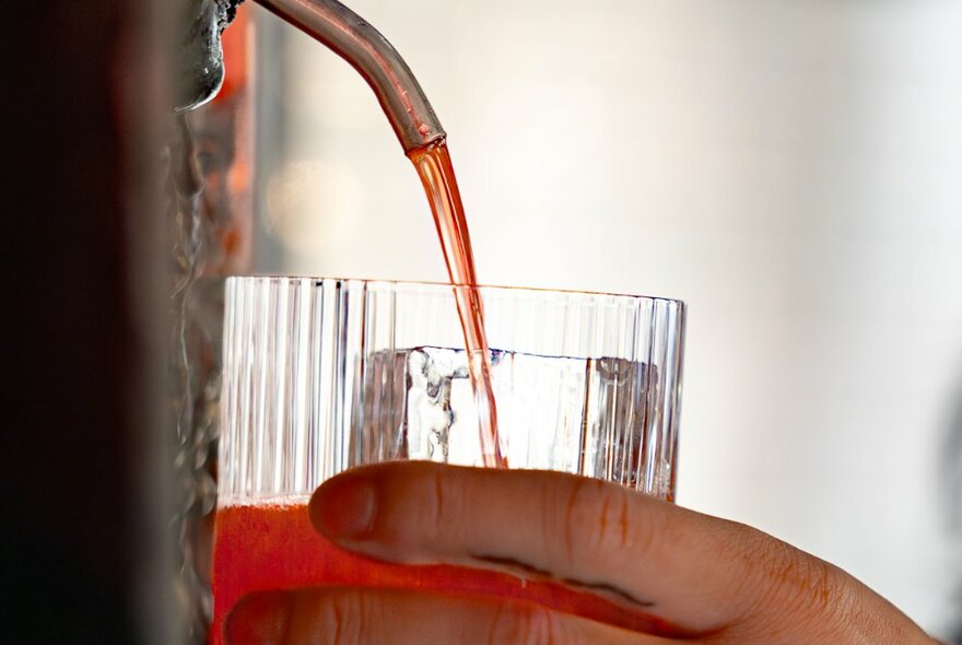 Hand holding a short glass under a mouth of a fountain dispensing negroni drinks