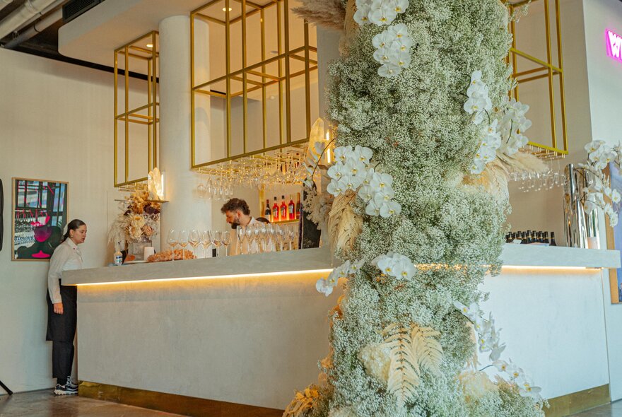 A cafe bar festooned with grey-green foliage and white blooms, staff behind the counter. 
