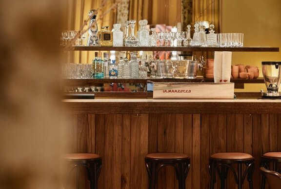 A wooden bar with a variety of glass and crystal stemware and a coffee machine, wooden stools in front of it.