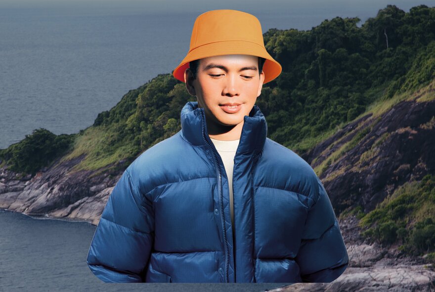  A man in an orange bucket hat and blue puffer jacket in front of a jagged coastline.