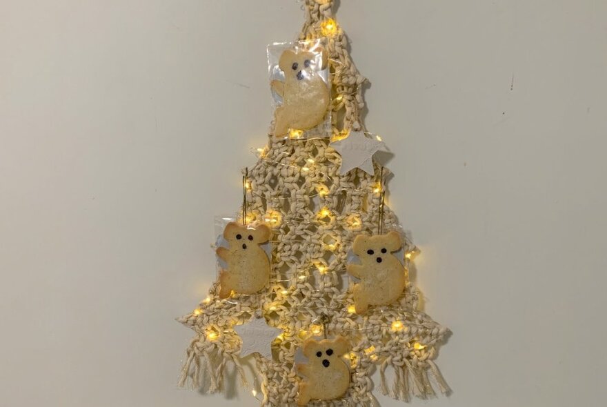 A flat macrame Christmas tree with small koala shaped decorations and fairy lights pinned to it, hanging on a wall.