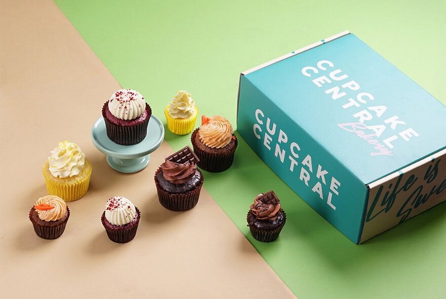 A selection of decorated cupcakes of different sizes, next to a takeaway green cardboard box with the words CUPCAKE CENTRAL written on the side.