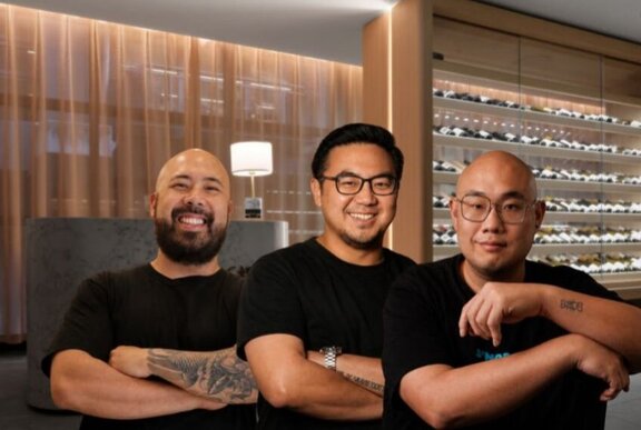 Three chefs wearing black t-shirts with arms crossed in front of their chest, behind them is a wall of wine bottles.