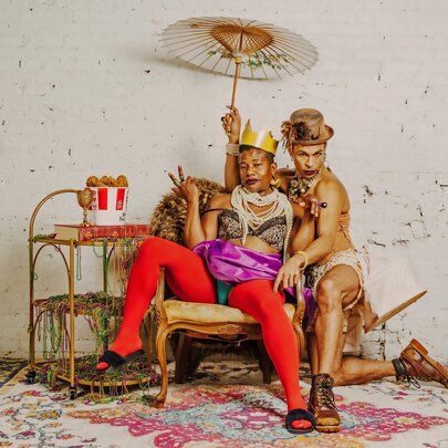 A person wearing a gold crown and bright red tights sitting on a vintage armchair, giving a peace sign. Another person in a top hat and a light-coloured outfit is sitting next to them, holding a white parasol over both of them. They are in a room with a white textured wall and a patterned rug.