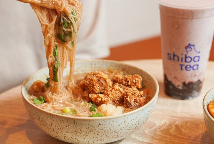 Chopsticks taking noodles from a bowl of soup next to a drink container of tea.