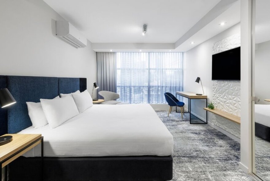 Hotel room with large bed facing a TV screen on the wall, with curtained windows at one end of the room and a writing nook.