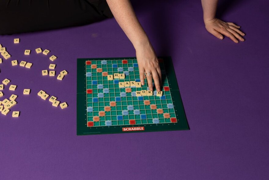A scrabble board on the floor, with an unseen person lying next to it, one hand holding them up and the other moving scrabble tiles on the board.