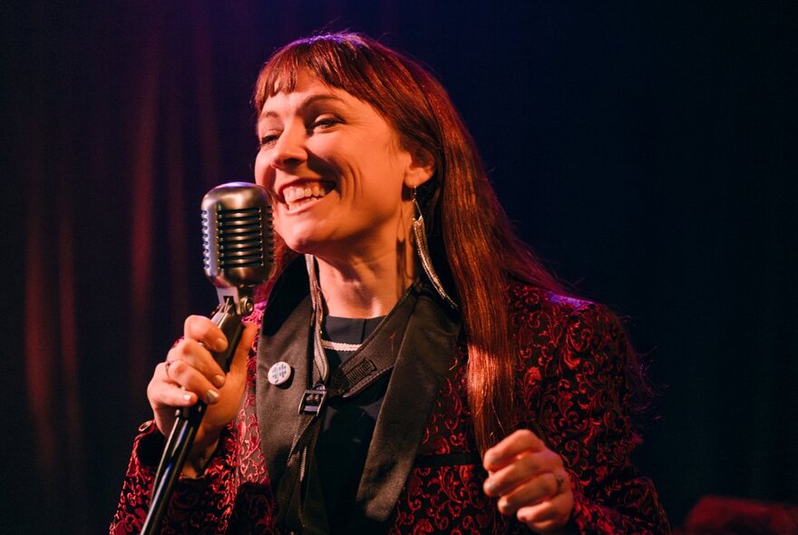 Singer Susy Hull smiling while holding a microphone on a dark stage.